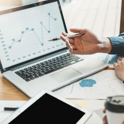 Die Hand einer Person zeigt auf einen geöffneten Laptop. Auf dem Bildschirm ist ein Liniendiagramm abgebildet und auf den Tisch liegen verschiedene Zettel und ein iPad.